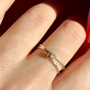 Op de afbeelding: Een goudkleurige ring met een uniek kruisbandontwerp en een klein, rond accent. De ring wordt op een vinger gedragen, met een warme, rood-oranje achtergrond.