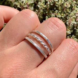 Könnte beinhalten: Drei silberne Ringe, die auf einem Finger übereinander liegen. Der obere Ring ist ein gehämmertes Band, der mittlere Ring ist ein gedrehtes Band und der untere Ring ist ein flaches Band.