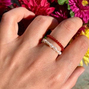 May include: A hand displaying two beaded rings. One ring is adorned with small, white, irregularly shaped pearls, and the other with small, red beads. The rings are set against a backdrop of vibrant, dark pink flowers.