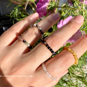 Op de afbeelding: Een hand toont vijf kralenringen in verschillende kleuren. De ringen zijn gemaakt van kleine, ronde kralen in zwart, groen, roze, geel en helder. De ringen bevinden zich op een hand met een bloemenachtergrond. Tekst op de afbeelding luidt: https://moonrubyshop.etsy.com.