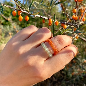 Op de afbeelding: Een hand met twee kralenringen. Eén ring is oranje, de andere lichtgroen. De ringen zijn gemaakt van kleine, ronde kralen. De achtergrond toont een tak met oranje bessen.