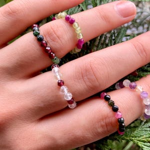 Könnte beinhalten: Ein Set aus fünf Perlenringen, jeder mit einer anderen Farbkombination. Die Ringe bestehen aus kleinen, runden Perlen in den Farben Weiß, Rosa, Grün, Schwarz und Gelb.