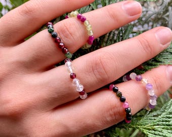 Könnte beinhalten: Ein Set aus fünf Perlenringen, jeder mit einer anderen Farbkombination. Die Ringe bestehen aus kleinen, runden Perlen in den Farben Weiß, Rosa, Grün, Schwarz und Gelb.