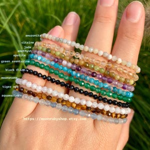 Könnte beinhalten: Eine Sammlung von 10 Edelstein-Armbändern, jedes mit einem anderen Stein. Die Armbänder sind übereinander gestapelt, mit dem Amazonit-Armband oben und dem Aquamarin-Armband unten. Die Armbänder sind alle aus kleinen, runden Perlen gefertigt. Die Steine umfassen Amazonit, Citrin, Jade, Amethyst, Apatit, grüner Aventurin, schwarzer Feuerstein, Mondstein, Tigerauge und Aquamarin.