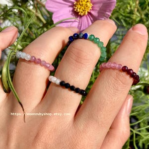 May include: Four beaded rings on a hand. The rings are made of different colored stones, including pink, green, blue, and black.
