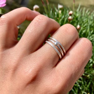 Könnte beinhalten: Ein Set aus drei gestapelten Silberringen an einem Finger. Das Set besteht aus zwei glatten, schlichten Silberringen und einem strukturierten Silberring mit einer kleinen, mittig platzierten Silberkugel. Das Metall hat ein mattes, gebürstetes Finish.
