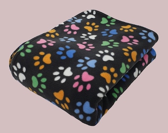 Könnte beinhalten: Gefaltete schwarze Fleece-Decke mit einem farbenfrohen Pfotenabdruckmuster. Die Pfotenabdrücke sind in den Farbtönen Rosa, Blau, Gelb, Grün und Weiß gehalten. Die Decke ist für Haustiere konzipiert.
