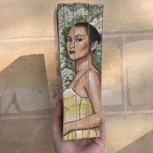 Pode incluir: Uma obra de arte pintada &agrave; m&atilde;o com um retrato de uma mulher com cabelo escuro e batom vermelho, vestindo um vestido amarelo. O fundo tem um design floral verde e branco. A obra de arte est&aacute; em uma superf&iacute;cie de madeira.