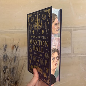Puede incluir: Un libro de tapa dura titulado "Maxton Hall Save Me" de Mona Kasten. La portada es azul oscuro con detalles dorados e ilustraciones. El borde del libro presenta retratos de dos personas. El libro está sostenido por una mano.