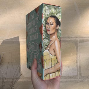 Pode incluir: Um livro intitulado "The Complete Summer I Turned Pretty Trilogy" de Jenny Han. A capa apresenta uma mulher com um vestido amarelo, em um fundo de flores brancas. A lombada &eacute; turquesa com ilustra&ccedil;&otilde;es de conchas.