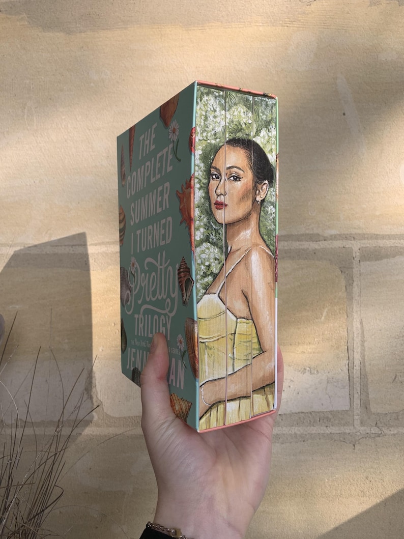 Pode incluir: Um livro intitulado "The Complete Summer I Turned Pretty Trilogy" de Jenny Han. A capa apresenta uma mulher com um vestido amarelo, em um fundo de flores brancas. A lombada &eacute; turquesa com ilustra&ccedil;&otilde;es de conchas.