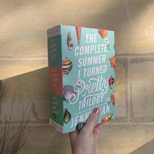 Pode incluir: Um conjunto de livros intitulado "The Complete Summer I Turned Pretty Trilogy" de Jenny Han. A capa do livro &eacute; azul claro com ilustra&ccedil;&otilde;es de conchas e o t&iacute;tulo em branco e rosa. O livro &eacute; segurado em uma m&atilde;o.