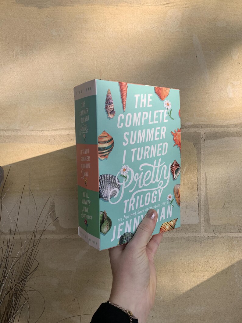 Pode incluir: Um conjunto de livros intitulado "The Complete Summer I Turned Pretty Trilogy" de Jenny Han. A capa do livro &eacute; azul claro com ilustra&ccedil;&otilde;es de conchas e o t&iacute;tulo em branco e rosa. O livro &eacute; segurado em uma m&atilde;o.