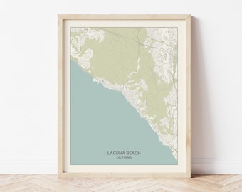 May include: A framed map of Laguna Beach, California, with a light blue background and green and brown lines representing roads and terrain. The map is in a light wood frame.