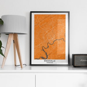 May include: A framed map of Knoxville, Tennessee, with orange and gray colors. The map is hanging on a white wall above a white shelf with a lamp, a speaker, and two plants.