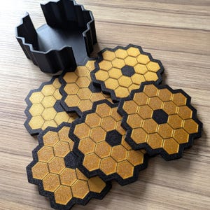 Puede incluir: Seis posavasos hexagonales negros y dorados con un patrón de panal. Los posavasos están apilados uno encima del otro. Un soporte para posavasos de plástico negro está en el fondo.