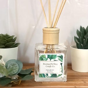 Może przedstawiać: Przezroczysty szklany dyfuzor patyczkowy z drewnianą nakrętką i patyczkami, oznaczony napisem "Boutique Na' Shay Candle Co." z zielonym wzorem liści. Dwa małe sukulenty w doniczkach flankują dyfuzor na drewnianej tacy.