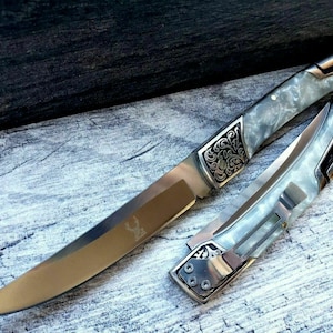 May include: A silver folding knife with a white, marbled handle. The handle is decorated with intricate floral designs. The blade is long and curved.