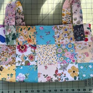 May include: A patchwork tote bag with floral patterns in various colors, including yellow, blue, pink, and white. The bag features two handles and a rectangular shape, with a mix of different fabric squares sewn together. The bag is on a green cutting mat.