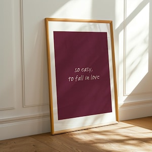 May include: A framed print with a burgundy background and the words "so easy, to fall in love" in white script. The frame is a light brown color, and the print is leaning against a white wall and wooden floor.