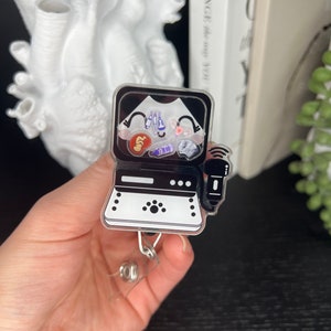 Pode incluir: Um porta-crachá de máquina de ultrassom em preto e branco com ilustrações médicas. O porta-crachá possui um cordão retrátil e um clipe de plástico transparente. As ilustrações incluem um feto, ferramentas médicas e um bebê.