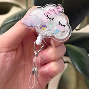 K&ouml;nnte beinhalten: Ein wei&szlig;er, wolkiger, einziehbarer Ausweishalter mit einem rosa und blauen Wolkenmuster, dem Text "ZZz" und einer Vielzahl kleiner, bunter Illustrationen. Der Halter ist an einem silbernen, einziehbaren Clip befestigt.