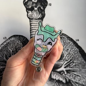 Peut inclure: Un porte-badge en plastique transparent avec une illustration de dessin animé d'un larynx humain. Le larynx est vert, rose et blanc avec un visage souriant et un œil qui cligne. Le porte-badge a un cordon rétractable.