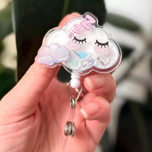 K&ouml;nnte beinhalten: Ein durchsichtiger Plastik-Badgehalter in Form einer Wolke mit einem schlafenden rosa und wei&szlig;en Gesicht. Die Wolke hat eine kleine wei&szlig;e Wolke im Inneren und ein rosa "zzz" &uuml;ber den Augen. Der Badgehalter ist an einer silbernen, einziehbaren Badge-Rolle befestigt.