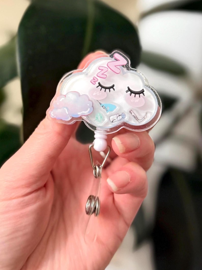 K&ouml;nnte beinhalten: Ein durchsichtiger Plastik-Badgehalter in Form einer Wolke mit einem schlafenden rosa und wei&szlig;en Gesicht. Die Wolke hat eine kleine wei&szlig;e Wolke im Inneren und ein rosa "zzz" &uuml;ber den Augen. Der Badgehalter ist an einer silbernen, einziehbaren Badge-Rolle befestigt.