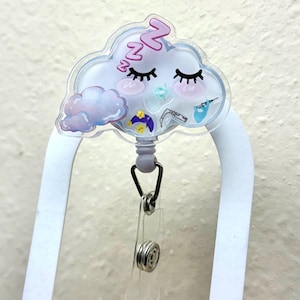 K&ouml;nnte beinhalten: Ein durchsichtiger Badgehalter aus Kunststoff mit einem schlafenden Wolkenmotiv. Die Wolke hat rosa Wangen, Wimpern und das Wort "zzz" darauf. Der Badgehalter hat eine einziehbare Schnur und einen Clip.