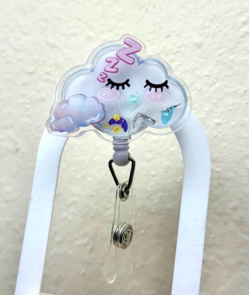 K&ouml;nnte beinhalten: Ein durchsichtiger Badgehalter aus Kunststoff mit einem schlafenden Wolkenmotiv. Die Wolke hat rosa Wangen, Wimpern und das Wort "zzz" darauf. Der Badgehalter hat eine einziehbare Schnur und einen Clip.