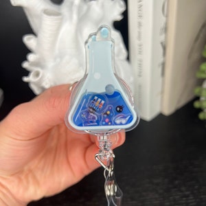 Può includere: Un porta badge retrattile a tema scientifico blu e bianco con un design a forma di becher in plastica trasparente. Il becher è riempito con icone colorate a tema scientifico e uno sfondo bianco.