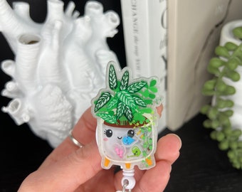 May include: A white plastic badge holder with a clear, round, resin charm featuring a potted plant with a cute face. The plant has green leaves and the pot has a white background with pink and blue details. The charm is attached to a white retractable badge reel.
