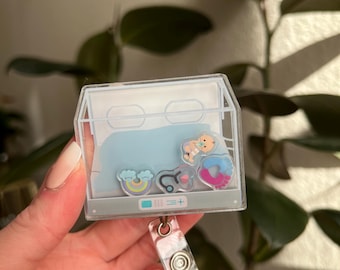 May include: A clear plastic badge holder with a blue background and various colorful cartoon images including a rainbow, a stethoscope, a baby's footprint, and a baby. The badge holder has a clear plastic retractable reel.