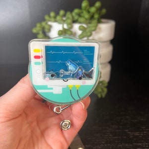 May include: A teal blue and white heart rate monitor badge reel with a cartoon ghost and a heart rate monitor graphic. The badge reel has a clear plastic cover and a silver clip.