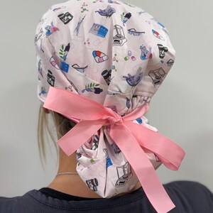 May include: A pink surgical scrub hat with a white background and a pattern of science-related illustrations, including beakers, test tubes, pills, and a scale. The hat has a pink ribbon tied at the back.