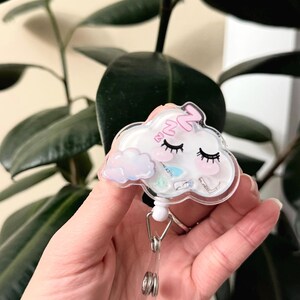 K&ouml;nnte beinhalten: Ein wei&szlig;er, wolkiger Badgehalter mit einem rosa schlafenden Gesicht und einer kleinen Wolke mit einem Regenbogen. Der Badgehalter hat eine transparente Plastikabdeckung und eine silberne, einziehbare Rolle.