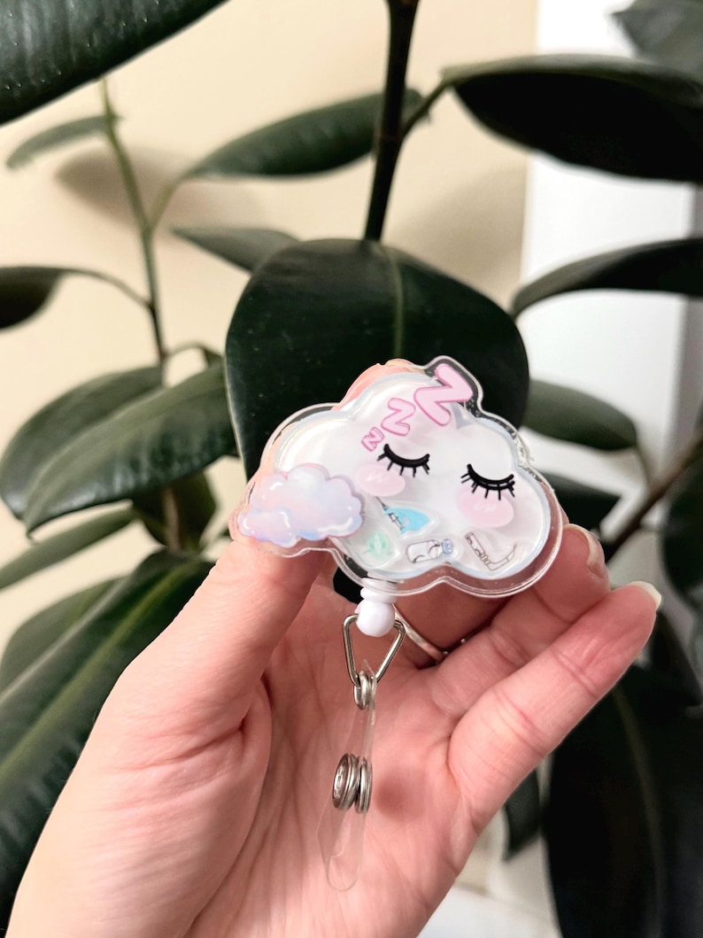 K&ouml;nnte beinhalten: Ein wei&szlig;er, wolkiger Badgehalter mit einem rosa schlafenden Gesicht und einer kleinen Wolke mit einem Regenbogen. Der Badgehalter hat eine transparente Plastikabdeckung und eine silberne, einziehbare Rolle.