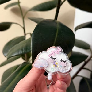 K&ouml;nnte beinhalten: Ein wei&szlig;er, wolkiger, einziehbarer Ausweishalter mit einem rosa und violetten schlafenden Wolken-Design. Die Wolke hat ein rosa Herz, einen Mond und Sterne. Der Halter hat eine wei&szlig;e, einziehbare Schnur und einen silbernen Clip.