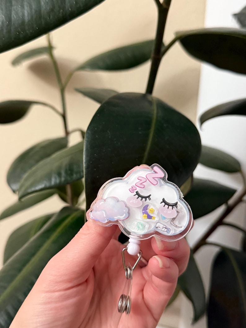 K&ouml;nnte beinhalten: Ein wei&szlig;er, wolkiger, einziehbarer Ausweishalter mit einem rosa und violetten schlafenden Wolken-Design. Die Wolke hat ein rosa Herz, einen Mond und Sterne. Der Halter hat eine wei&szlig;e, einziehbare Schnur und einen silbernen Clip.
