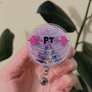May include: A clear plastic badge holder with a pink exercise ball design. The badge holder has a retractable cord and features a pink barbell with the letters "PT" in black.