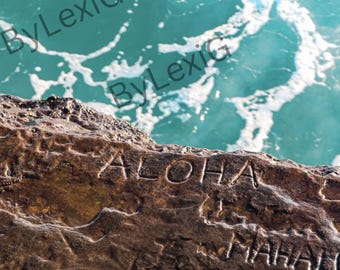 Puede incluir: Primer plano de una formación rocosa marrón con la palabra "ALOHA" grabada. El océano es visible en el fondo, con agua turquesa y olas blancas. La palabra "MAHALO" también está grabada en la roca.