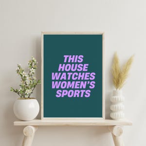 Pode incluir: Uma impress&atilde;o emoldurada em azul-petr&oacute;leo com o texto "THIS HOUSE WATCHES WOMEN'S SPORTS" em roxo sobre um fundo branco. A impress&atilde;o &eacute; exibida em uma prateleira de madeira com um vaso de flores brancas e um vaso de grama seca.