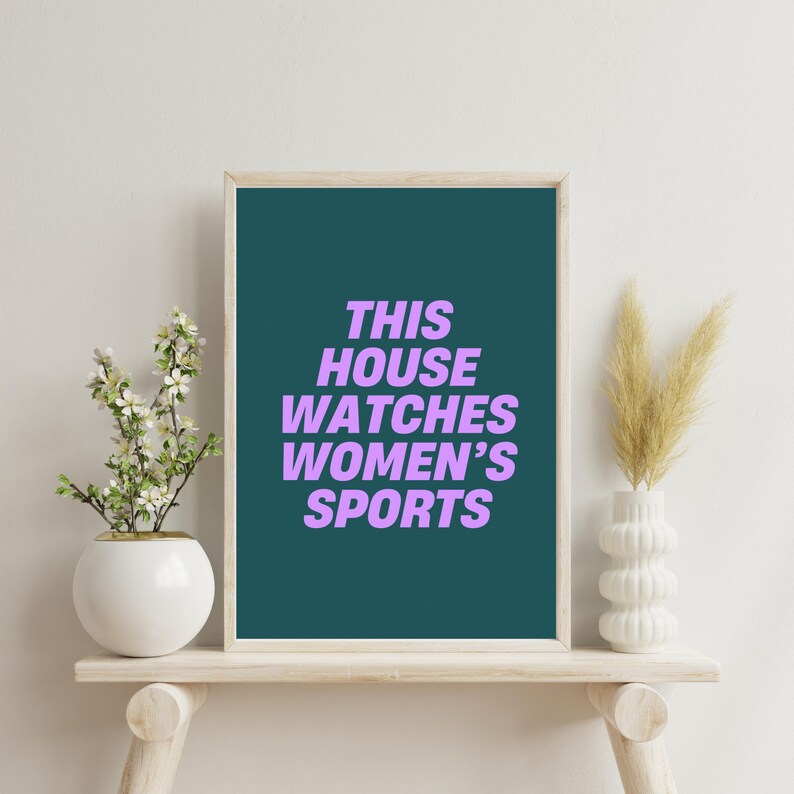 Pode incluir: Uma impress&atilde;o emoldurada em azul-petr&oacute;leo com o texto "THIS HOUSE WATCHES WOMEN'S SPORTS" em roxo sobre um fundo branco. A impress&atilde;o &eacute; exibida em uma prateleira de madeira com um vaso de flores brancas e um vaso de grama seca.