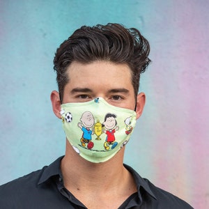May include: A light green face mask featuring a cartoon design of Charlie Brown and friends. The mask has a repeating pattern of soccer balls and characters. The mask is worn by a person wearing a black shirt.