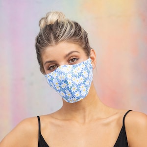 May include: A woman wearing a blue and white floral face mask with a white background and small blue flowers. The mask covers her nose and mouth. She is wearing a black tank top.