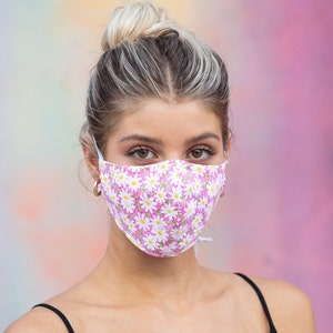 May include: A woman wearing a pink face mask with a white daisy pattern. The mask has a white elastic band.