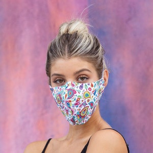 May include: A woman wearing a white face mask with a colorful paisley pattern. The mask covers her nose and mouth. She has blonde hair pulled back in a bun.