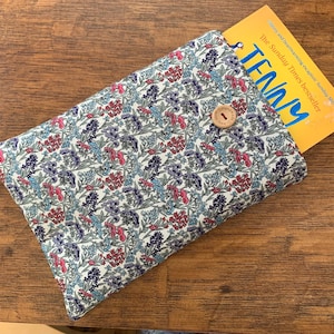 Può includere: Una custodia per libri in tessuto floreale, chiusa con un bottone di legno. La custodia contiene un libro con il titolo "TENNY". Il tessuto presenta un motivo di fiori rossi, blu e viola su sfondo crema.