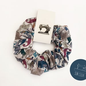 May include: A white scrunchie with a colorful animal print design. The scrunchie is tied in a knot and has a small, white tag with a black sewing machine graphic and the text "Sew Celia".